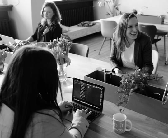 Three women at a desk, one working on a laptop.